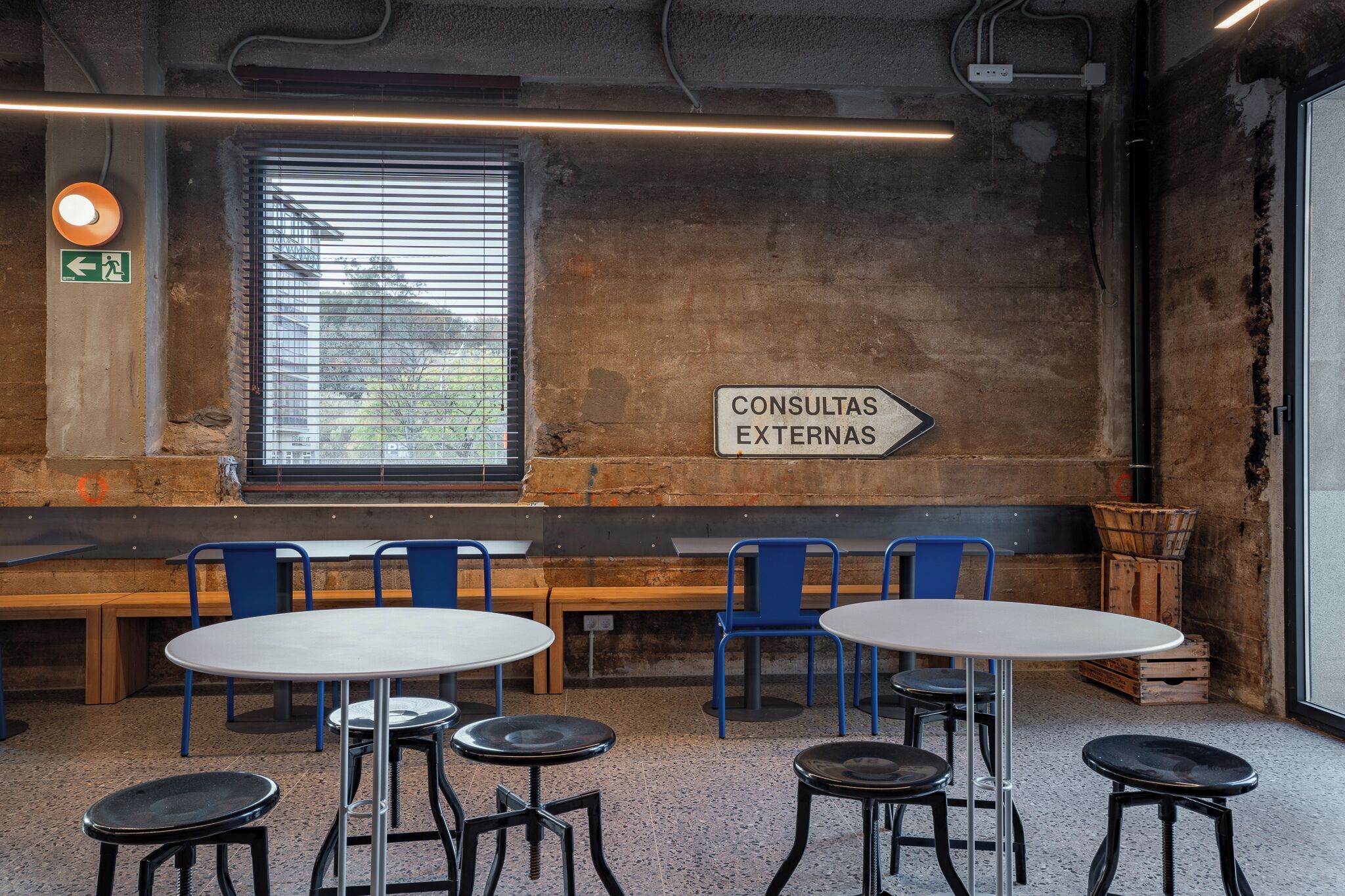 A rustic interior with a long bench, round tables, and blue chairs. A sign on the wall reads "CONSULTAS EXTERNAS".