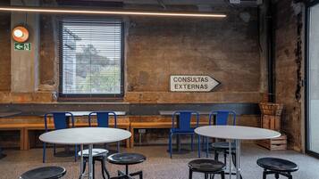 A rustic interior with a long bench, round tables, and blue chairs. A sign on the wall reads "CONSULTAS EXTERNAS".