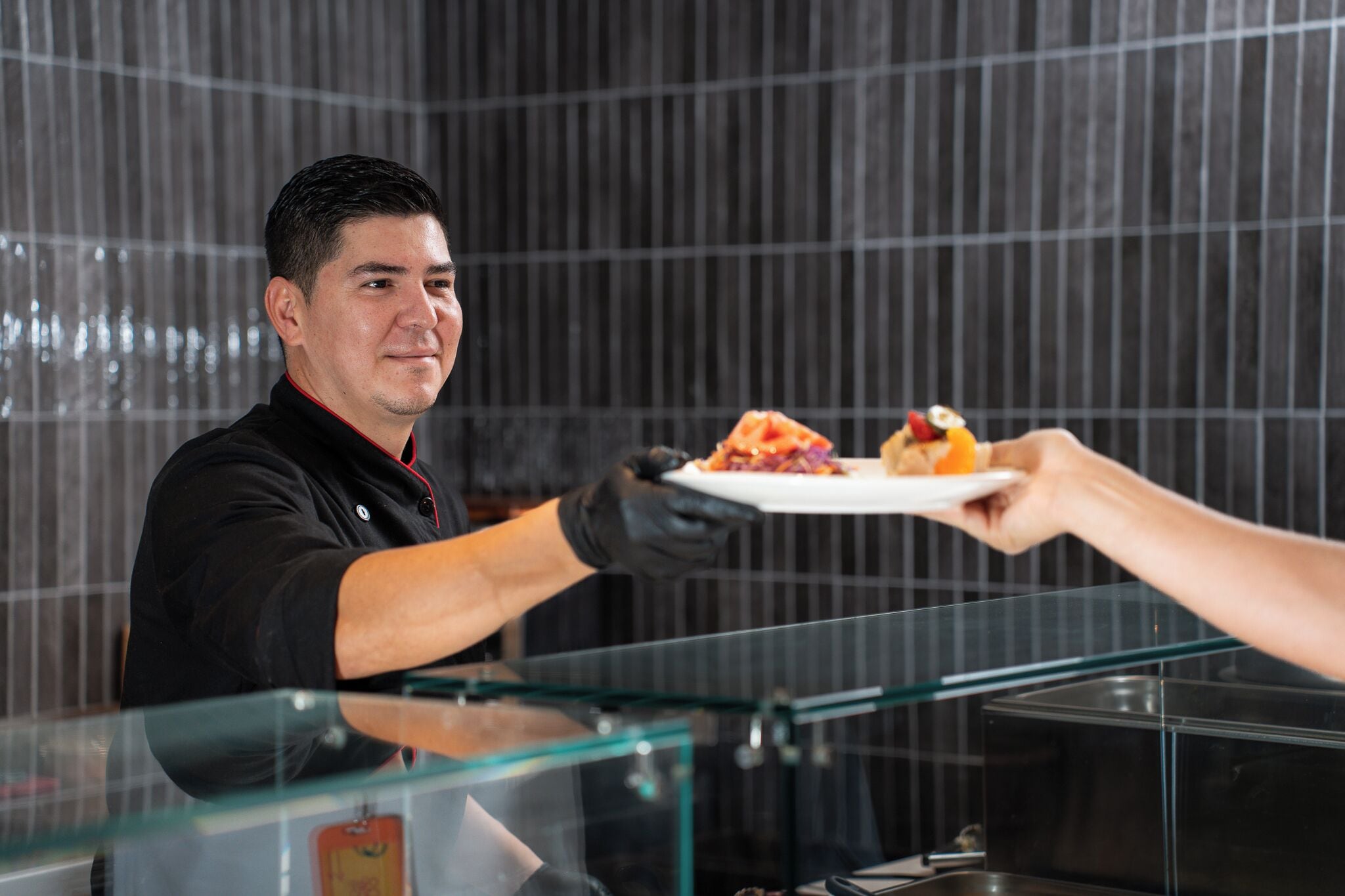Une personne servant des plats derrière un comptoir en verre, dans un décor moderne avec un mur à rayures en arrière-plan.
