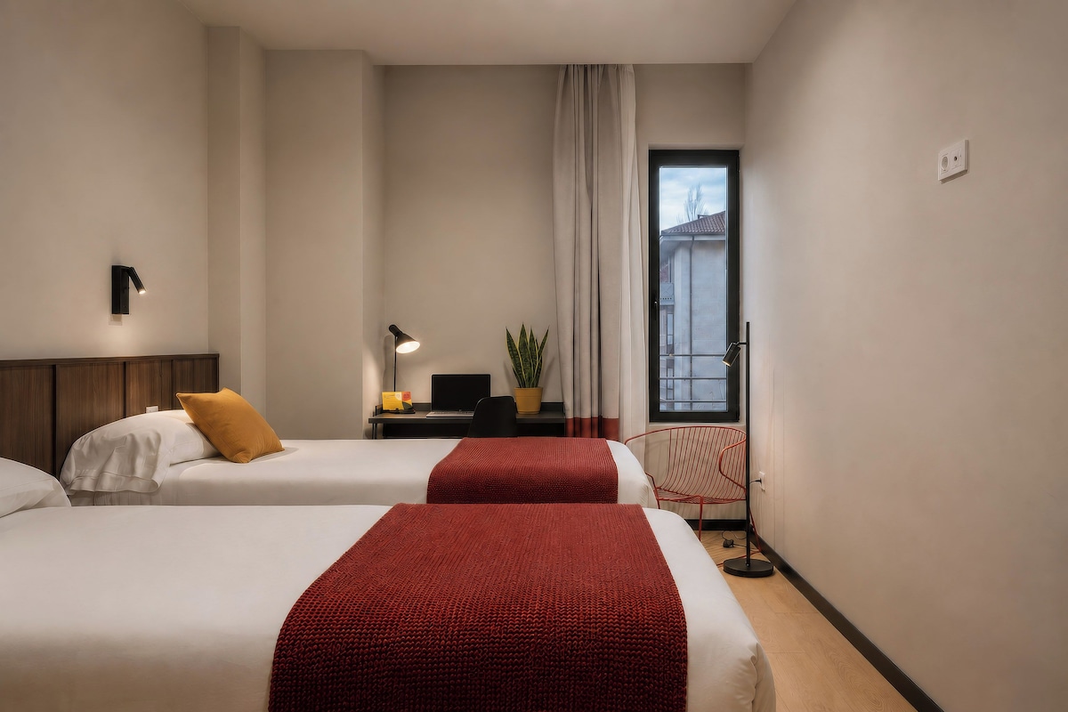 Habitación de hotel con dos camas, un escritorio con una computadora portátil, una planta en maceta, una silla y una ventana con vistas.
