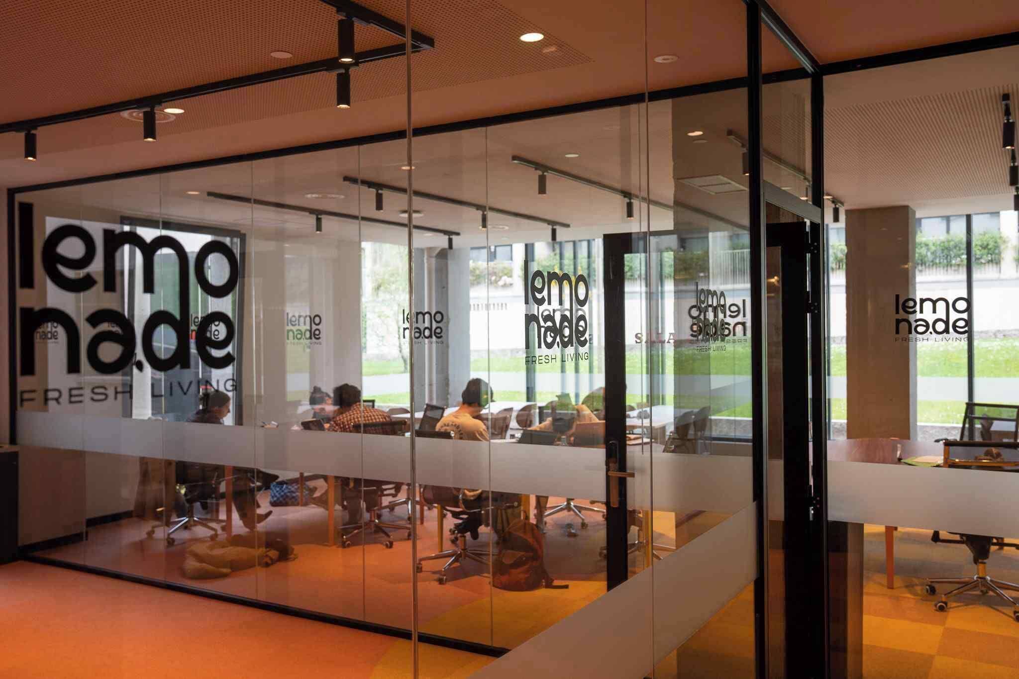 A modern office space with glass walls, multiple workstations, and the logo 'lemoade FRESH LIVING' on the glass.