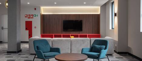 A modern lobby with a round wooden table, two blue armchairs, a red wall with a flat-screen TV, and a patterned floor.