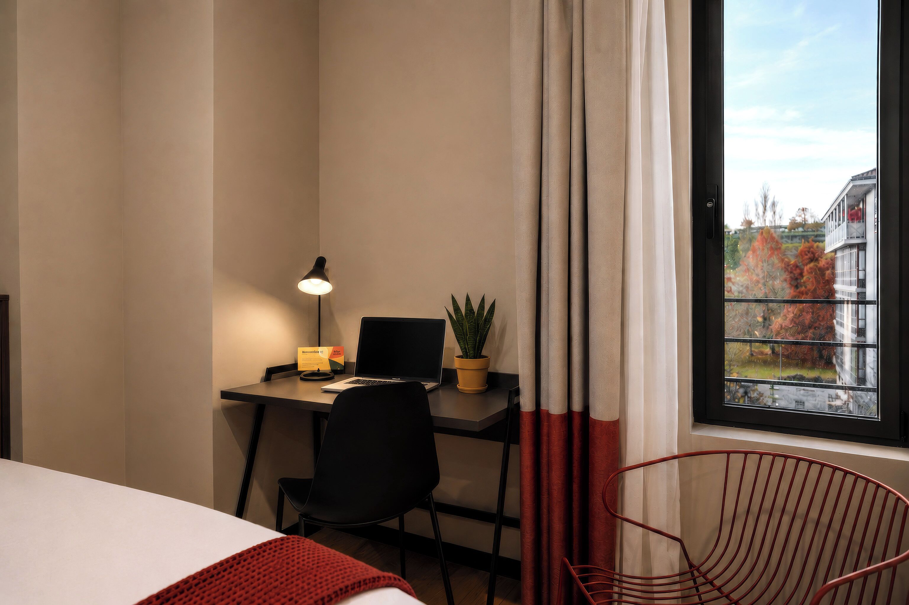A hotel room with a bed, a desk with a laptop, a chair, a potted plant, and a window with curtains.