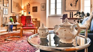 Un salon chaleureux avec un fauteuil rouge, une table basse en verre ornée de théières et une grande fenêtre donnant sur des œuvres d’art.