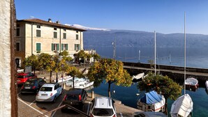 Un port de plaisance où sont garées des voitures et des bateaux, un bâtiment aux volets verts et une vue dégagée sur l’eau et les montagnes lointaines.