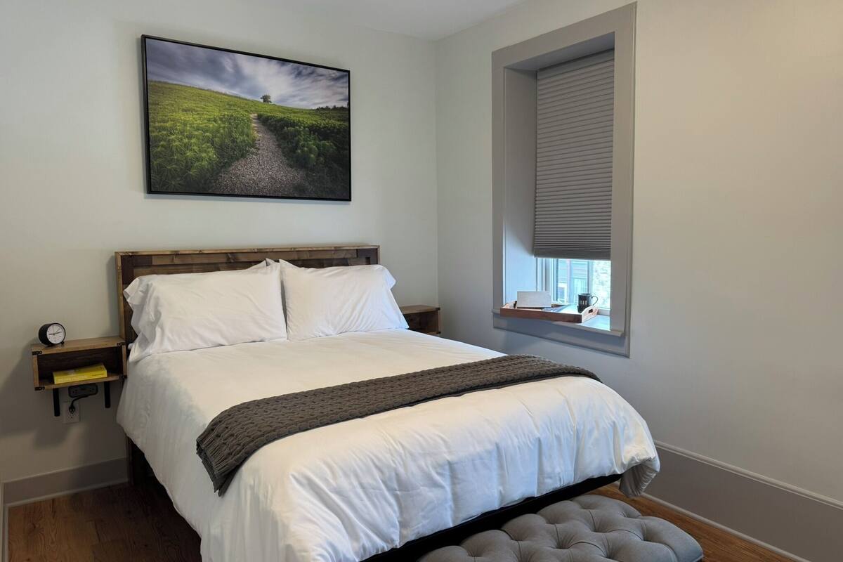 Un dormitorio con una cama grande, una mesita de noche, una ventana con persianas y un cuadro de paisaje en la pared.