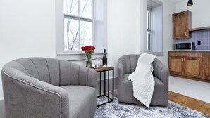A modern living room with two grey armchairs, a small side table with a vase of red flowers and a bottle, and a view of a kitchen with wooden cabinets and a microwave. - Brandt House (Alma)
