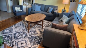 A living room with a sofa, armchair, coffee table, and patterned rug. There are framed pictures on the wall, a lamp, and a window with blinds.