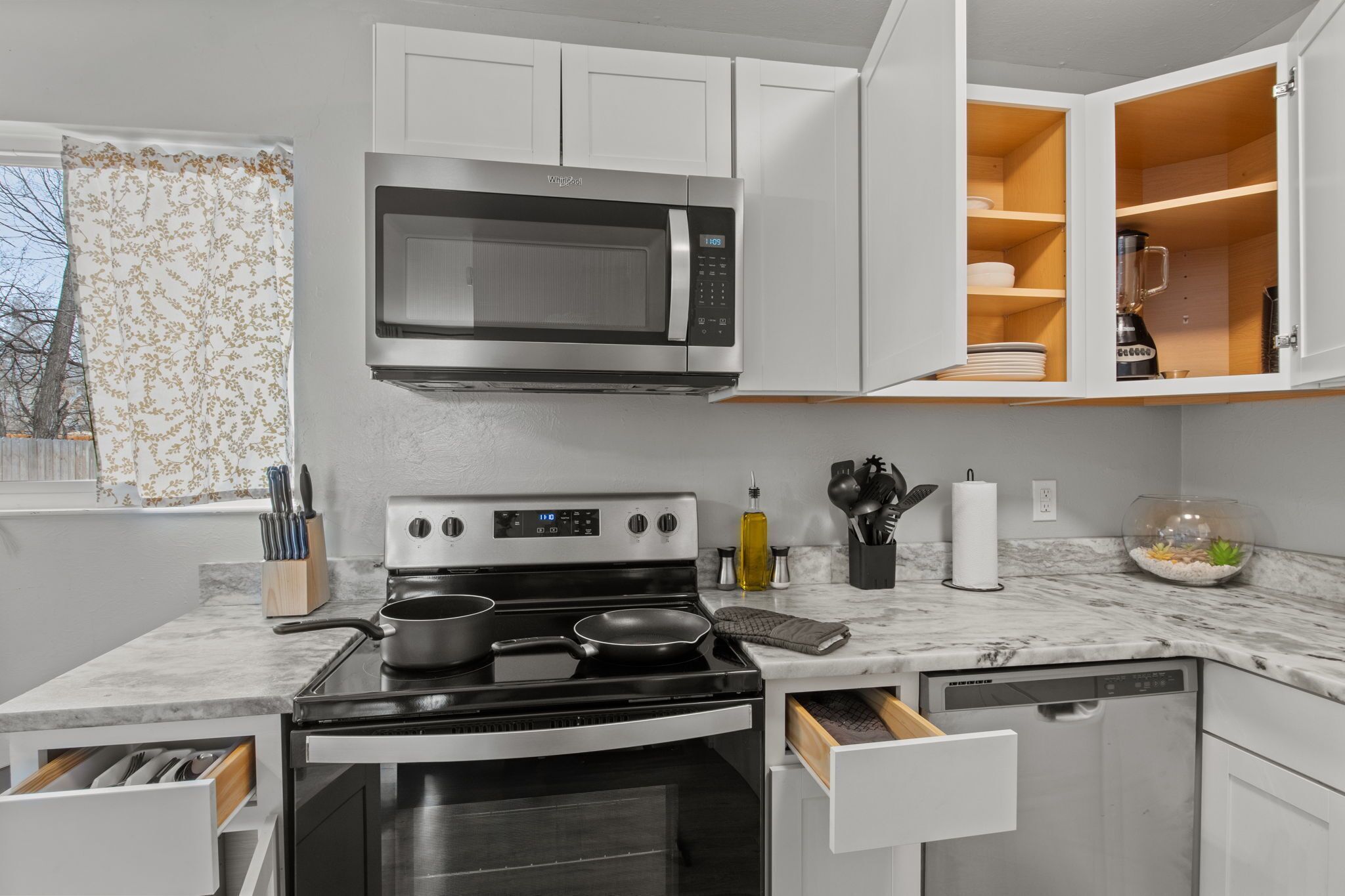 Une cuisine moderne équipée d’un four à micro-ondes, d’une cuisinière et d’un lave-vaisselle, avec des meubles blancs et une fenêtre agrémentée d’un rideau.