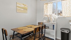 A dining area with a wooden table, four chairs, a white shelf with books, a vase with white flowers, and a window with curtains. - Family-Friendly w/ Hot Tub | 12 min to Broadmoor (Colorado Springs)