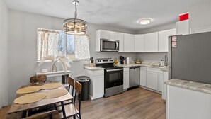 A modern kitchen with white cabinets, stainless steel appliances, a wooden dining table, and a hanging light fixture. - Family-Friendly w/ Hot Tub | 12 min to Broadmoor (Colorado Springs)