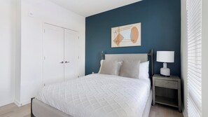 A bedroom with a bed, a nightstand, a lamp, a painting, and a door. - Landing | Stylish 1BD, Pool, Gym (Durham)