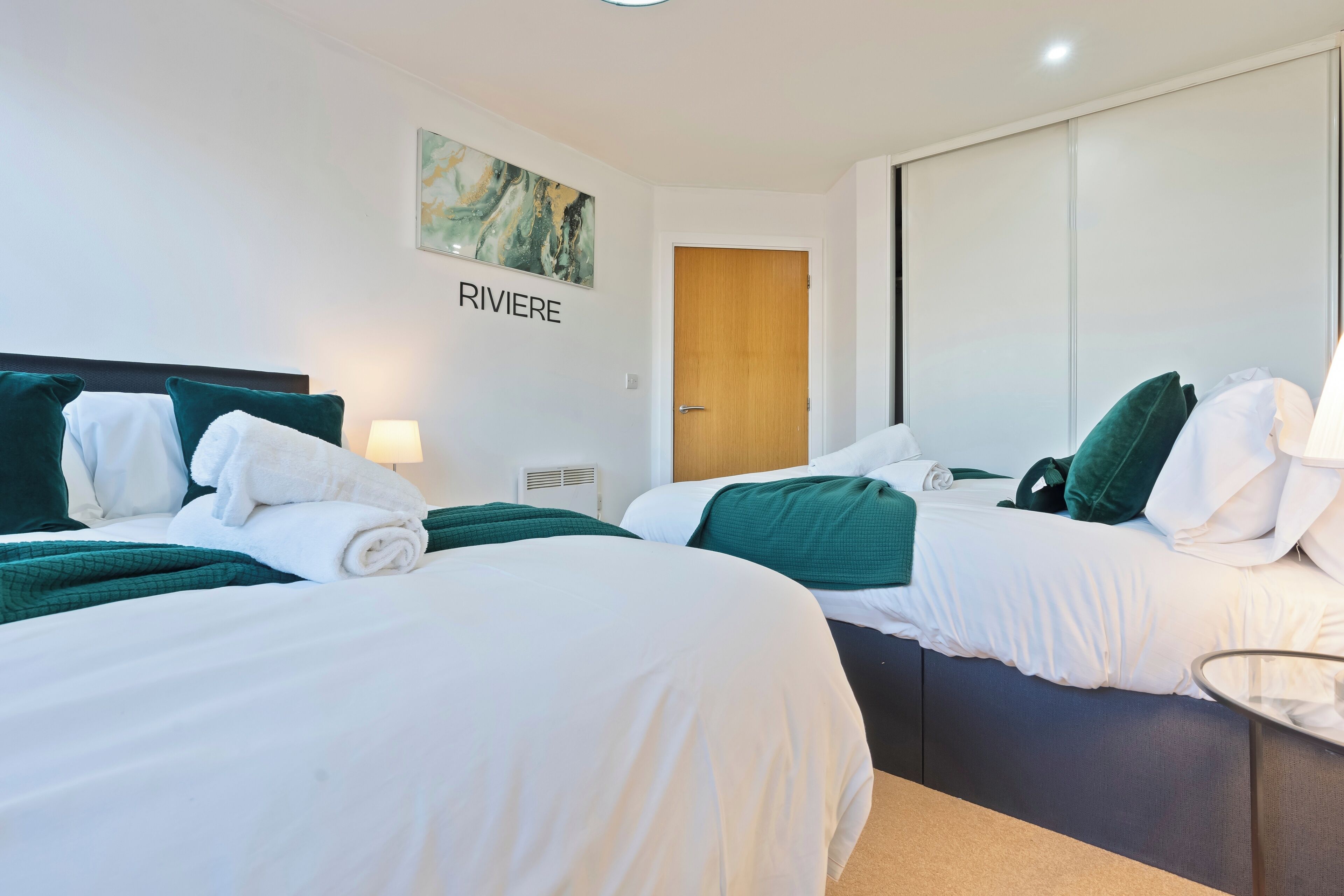 Une chambre d’hôtel avec deux lits, chacun étant agrémenté de draps verts et d’oreillers blancs. Un tableau est accroché au mur au-dessus du lit situé à gauche, et une porte se trouve à droite.