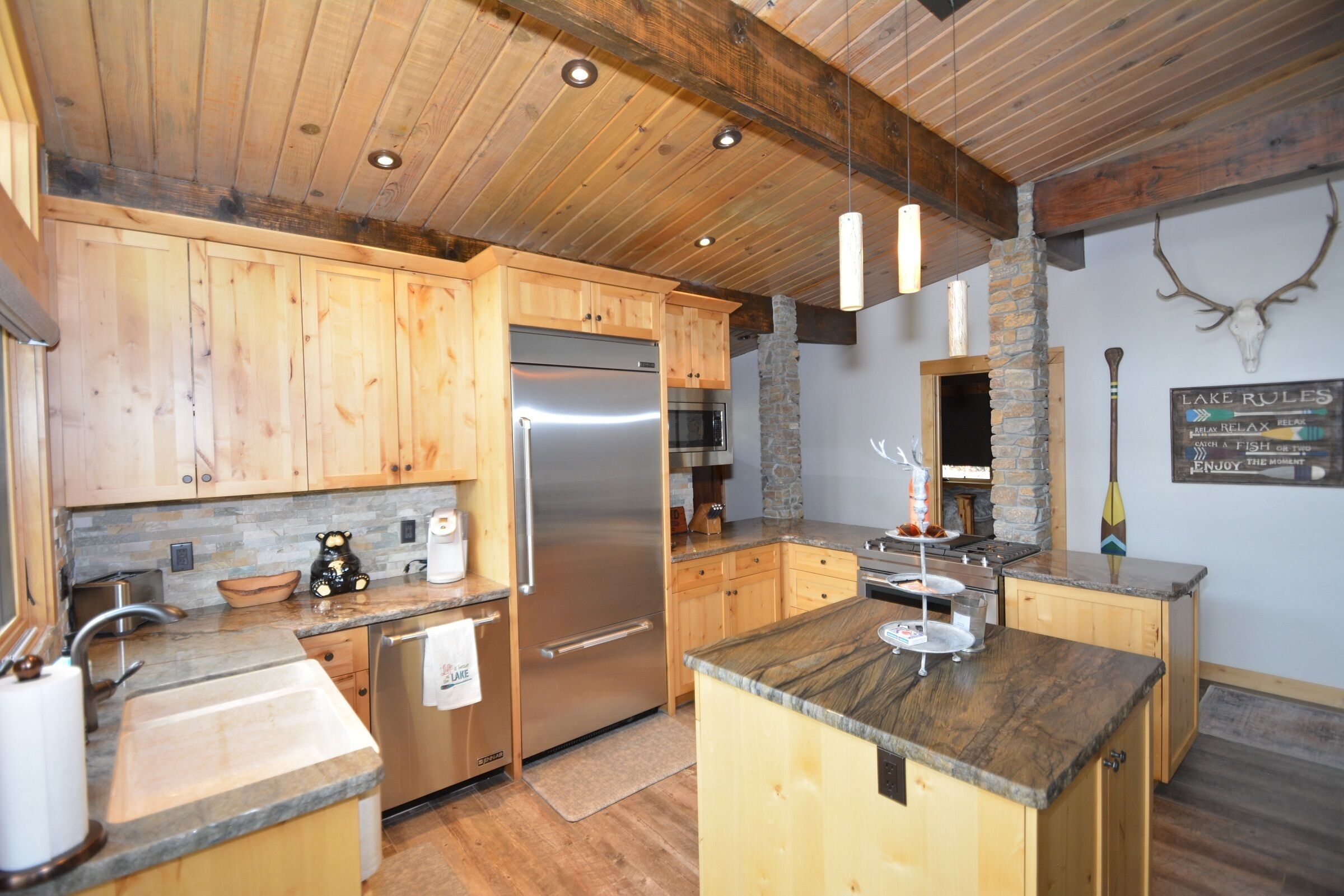 A modern kitchen with wooden cabinets, a stainless steel refrigerator, granite countertops, and a stone fireplace.