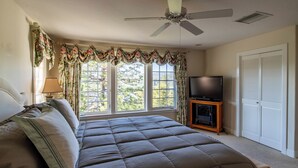 Un dormitorio con una cama grande, un ventilador de techo, un televisor sobre un soporte de madera y una ventana con cortinas.