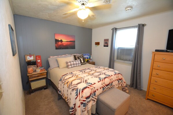 A bedroom with a bed, a nightstand, a dresser, a ceiling fan, a window with curtains, and a wall-mounted TV.