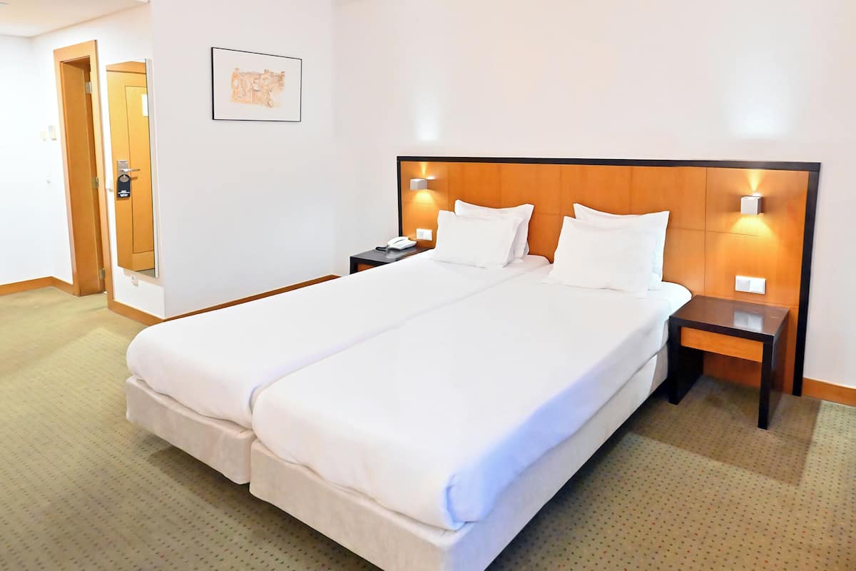 A hotel room with a large bed, two bedside tables, a nightstand, a wall-mounted light, and a door leading to another room.