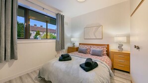 A bedroom with a bed, two bedside tables, a window with curtains, a wall-mounted light fixture, and a door with a handle. - Craigievar Crescent Apartment | Grampian Lettings (Aberdeen)