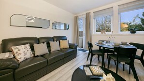 A modern living room with a black leather sofa, a dining table with chairs, and a large window with a view of the outdoors. - Craigievar Crescent Apartment | Grampian Lettings (Aberdeen)