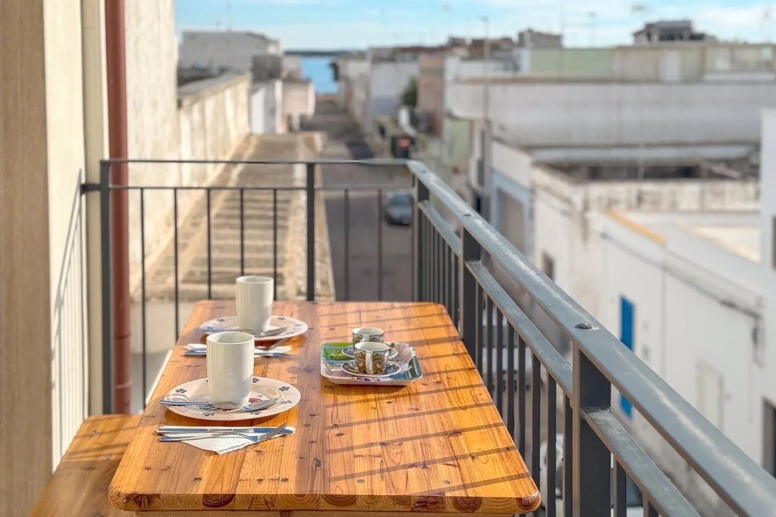 A balcony with a wooden table set for two, including plates, cups, and cutlery.