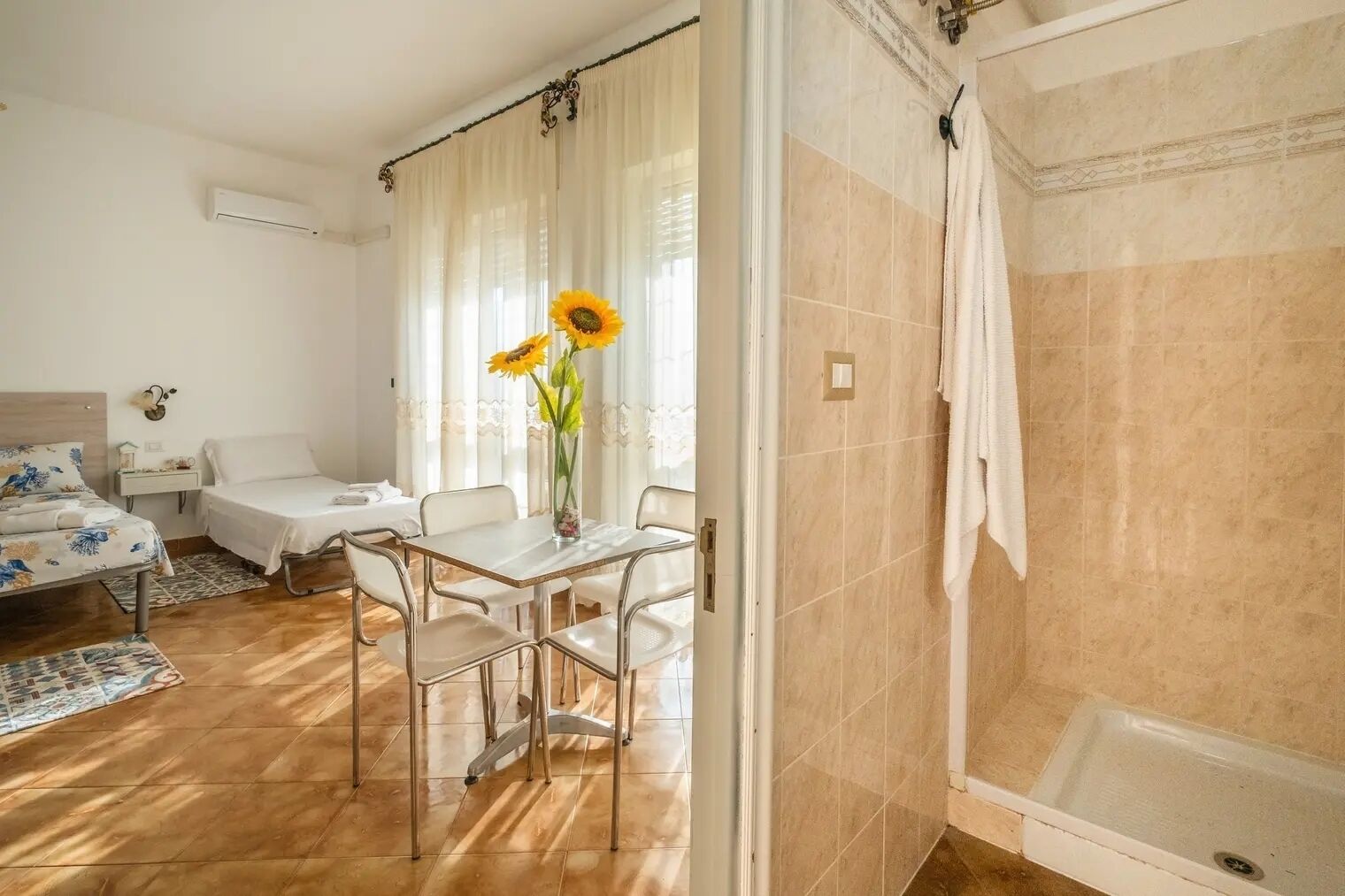 A hotel room with a bed, a small table with chairs, a vase with sunflowers, a shower area, and a window with curtains.
