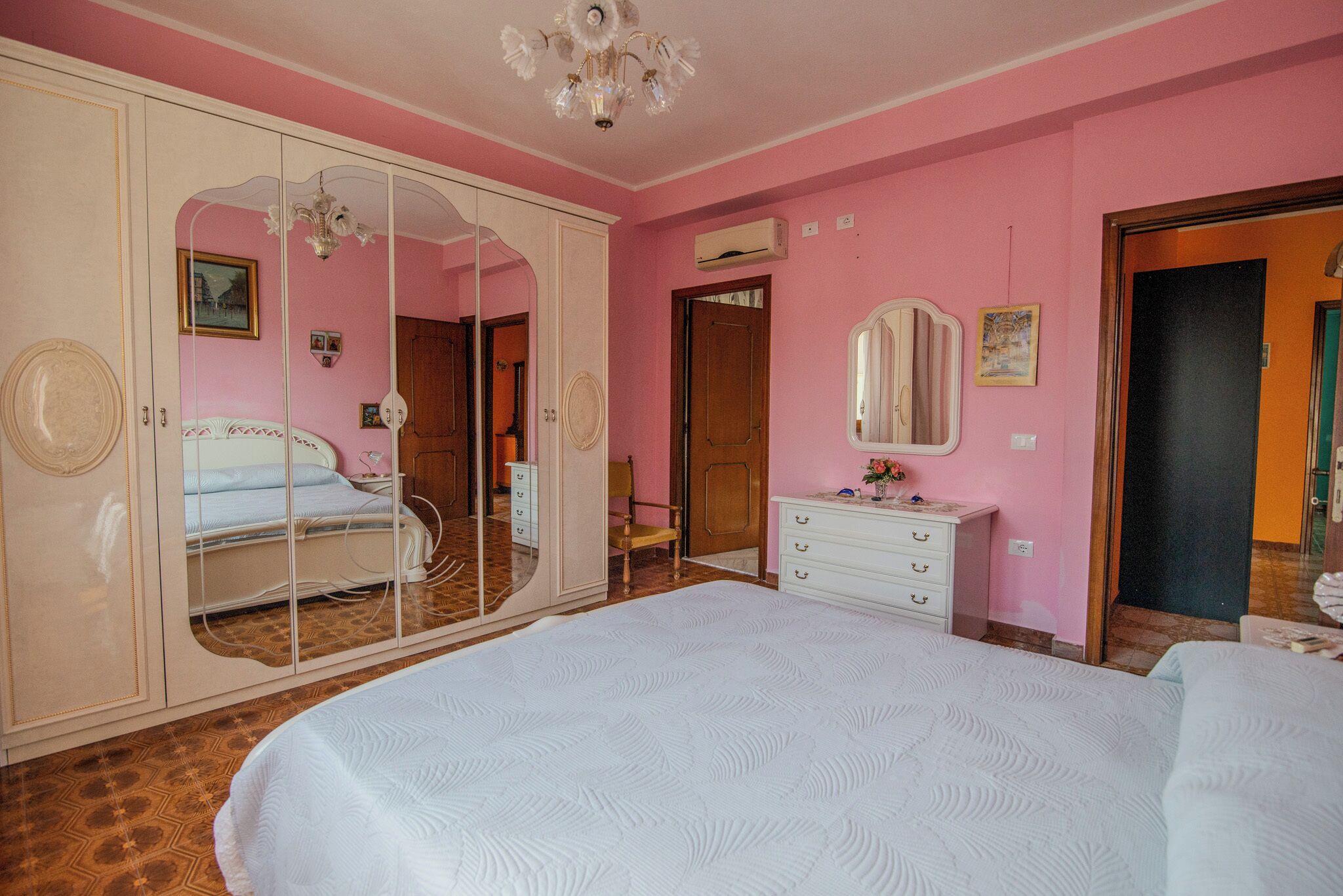 A bedroom with a large bed, a wardrobe with a mirror, a dresser, and a door leading to another room.