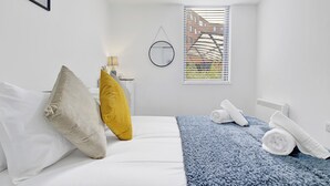 A hotel room with a bed, pillows, a blue blanket, a window with blinds, and a radiator. - Cosy Birmingham Apartment - Madison House (Birmingham)