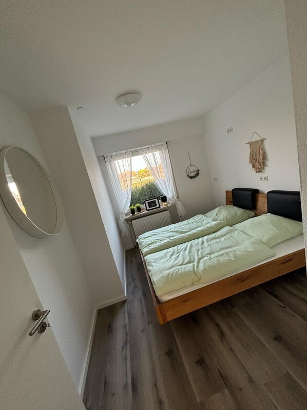 Un dormitorio con cama de madera, una ventana con cortinas translúcidas, un espejo montado en la pared y un tapiz de macramé.