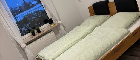 A bedroom with a wooden bed, white walls, a window with a view of a snowy landscape, and a radiator below the window.