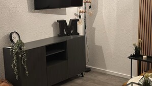 Una sala de estar moderna con un televisor de pantalla plana montado en la pared, un mueble de entretenimiento de color oscuro con un reloj y plantas, y una mesa de centro de vidrio con un control remoto.