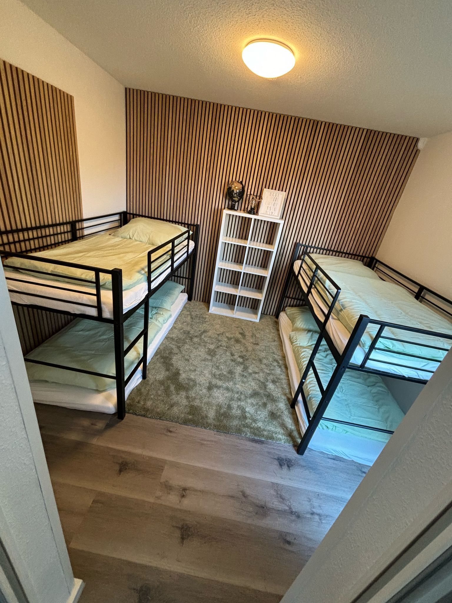 A dormitory room with bunk beds, a small white shelf with items, and a sloped ceiling with a light fixture.
