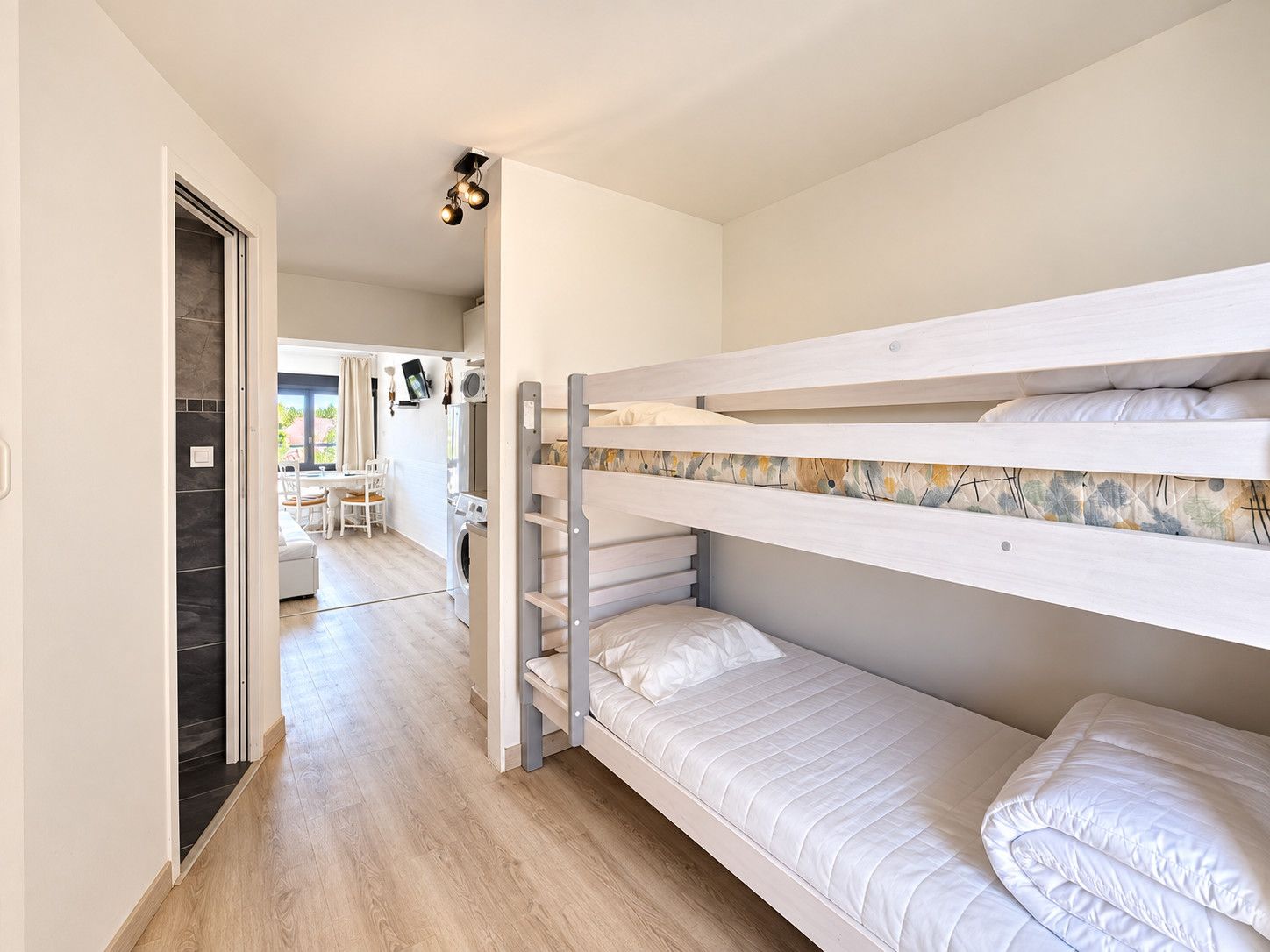 Una cama litera con estructura de madera y ropa de cama blanca, un pasillo con una puerta que da a un comedor, y una lámpara en el techo.