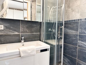 Ein modernes Badezimmer mit einer gläsernen Duschkabine, einem weißen Waschbecken und dunkel gefliesten Wänden.