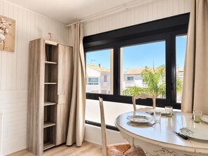 Ein Zimmer mit Esstisch für zwei Personen, einem Fenster mit Blick auf Gebäude und einem Holzregal.