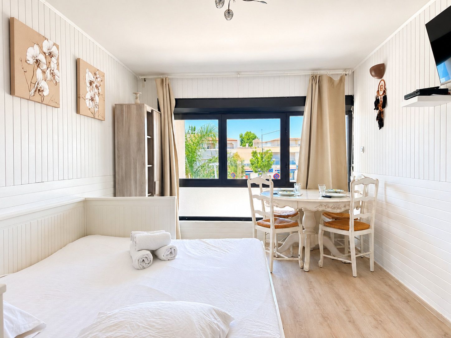 Una habitación de hotel con cama, una mesa de comedor con sillas, ventana con cortinas y obras de arte en las paredes.
