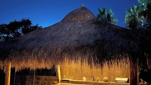 Una zona d'exterior per dinar, amb una estructura de palla, taula de fusta i un mur de pedra al fons.