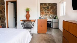 Ein Schlafzimmer mit Bett, Sessel, Schreibtisch, einer Steinwand, einer Küchenzeile mit Kühlschrank und einem an der Wand montierten Fernseher.