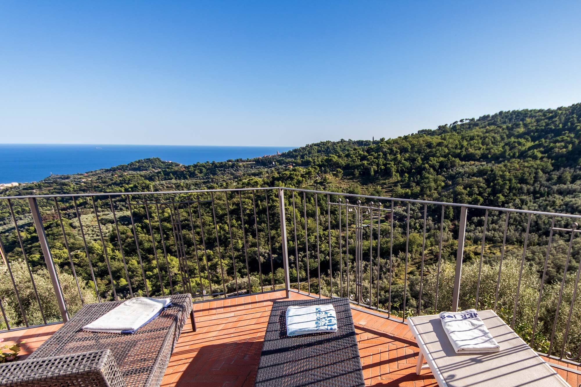 A terrace with two sun loungers and a view of the sea and forested hills.