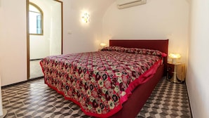 A bedroom with a patterned bedspread, a red headboard, a nightstand with a lamp, and a wall-mounted air conditioner. - Large panoramic sea view terrace, stone shower, excellence vacation. (Lingueglietta)
