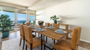 A dining area with a wooden table set for four, surrounded by leather chairs, with a view of the sea through a glass door.