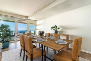 A dining area with a wooden table set for four, surrounded by leather chairs, with a view of the sea through a glass door.