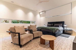 A modern bedroom with a bed, two armchairs, a small table, and a ceiling fan.