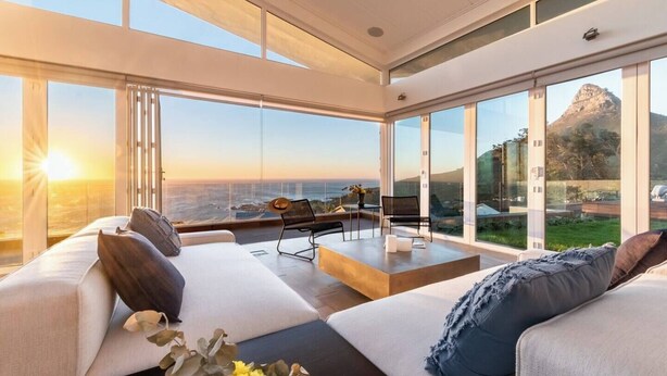 A modern living room with a large glass wall offering a view of the ocean and a mountain. There is a sofa set and a small table with chairs.