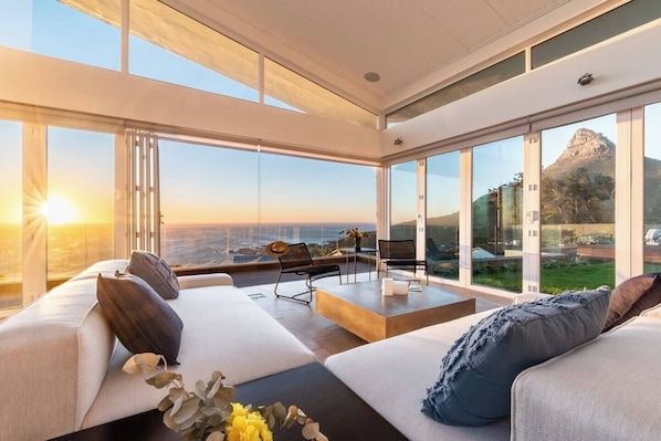 A modern living room with a large glass wall offering a view of the ocean and a mountain. There is a sofa set and a small table with chairs.