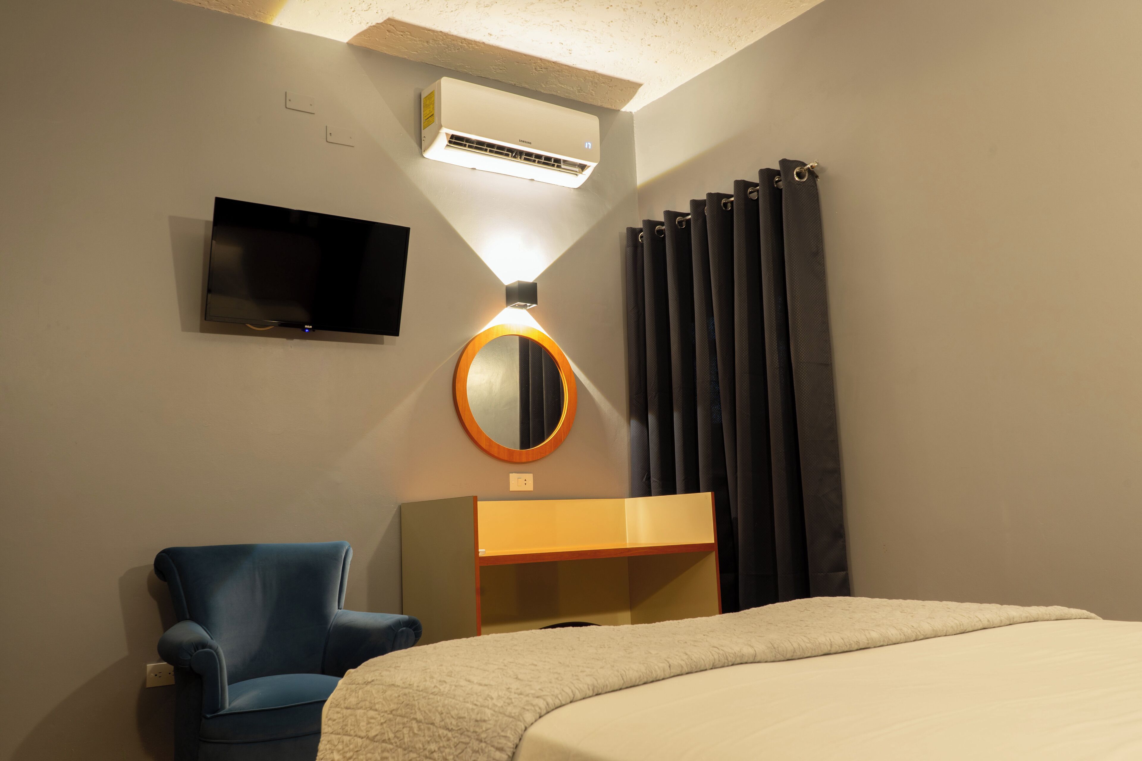 Habitación de hotel con cama, sillón azul, escritorio, televisión, aire acondicionado, espejo y cortinas.