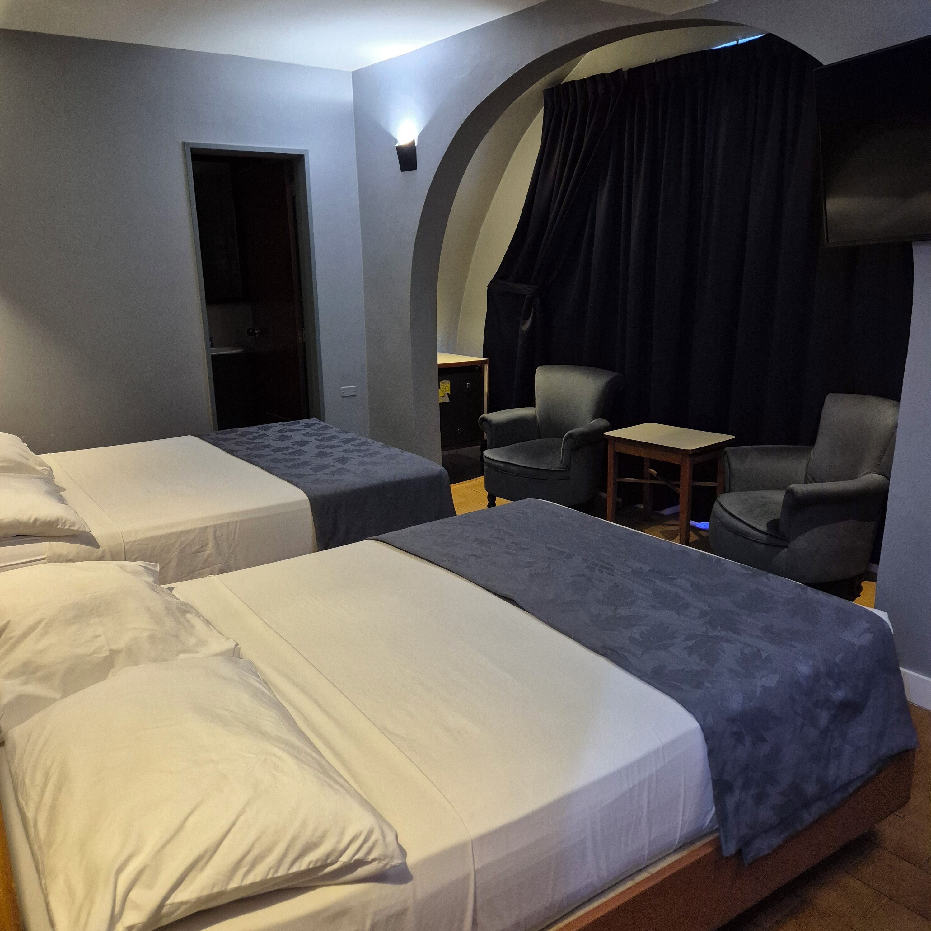 A hotel room with two beds, a small table, a chair, and a dark curtain.
