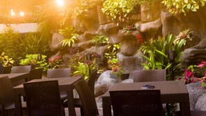 An outdoor dining area with wicker tables and chairs, surrounded by a variety of potted plants and a rock wall. - Hotel El Arroyo (Caracas)