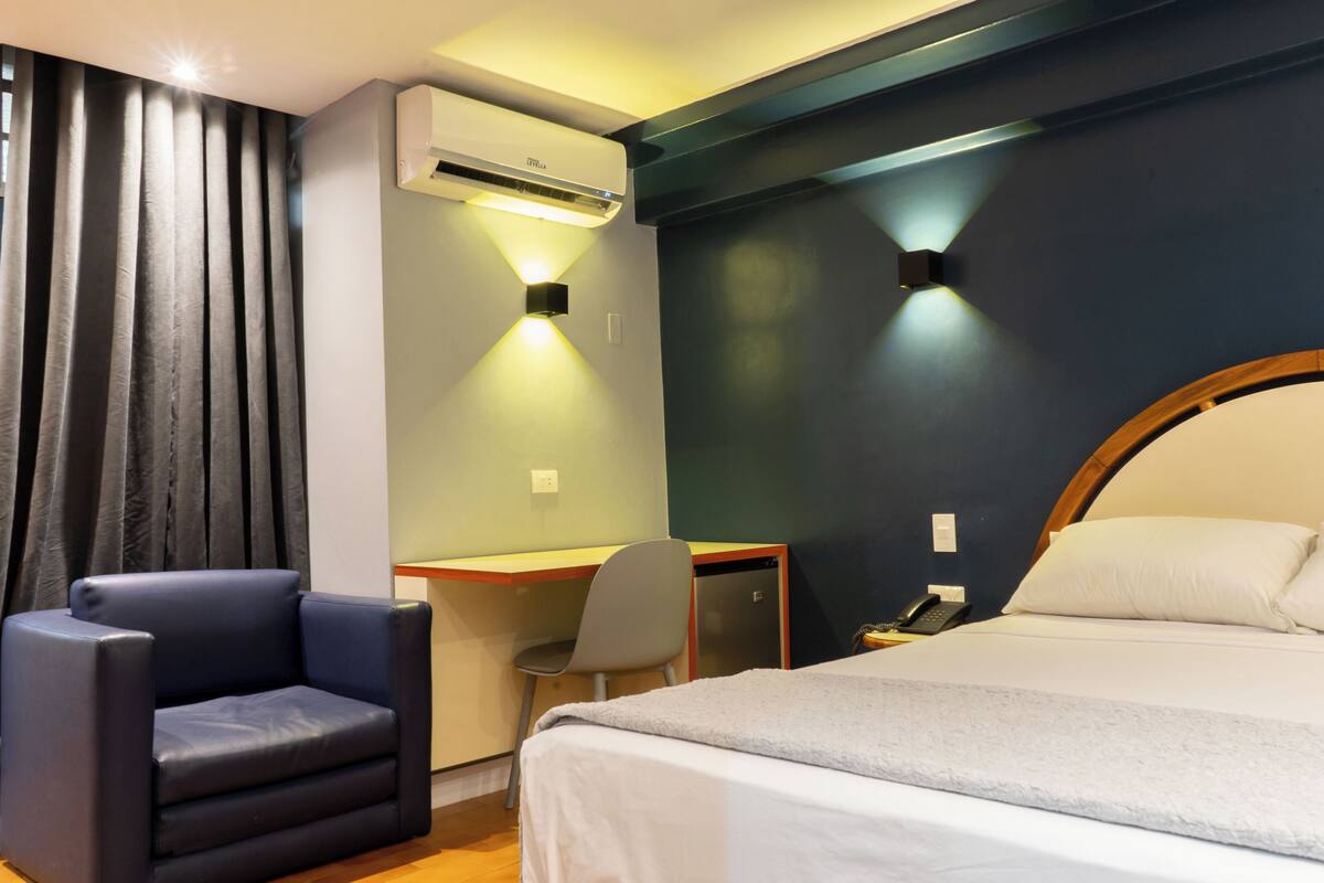 Een hotelkamer met een bed, een bureau, een stoel, een airconditioner en een telefoon aan de muur.
