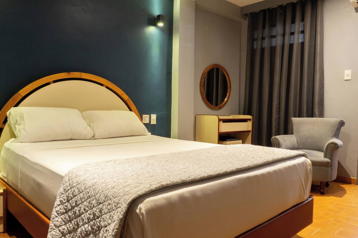 Een net opgemaakt bed met houten hoofdeinde, een nachtkastje en een stoel in een kamer met een donkere wand en een raam met gordijnen.