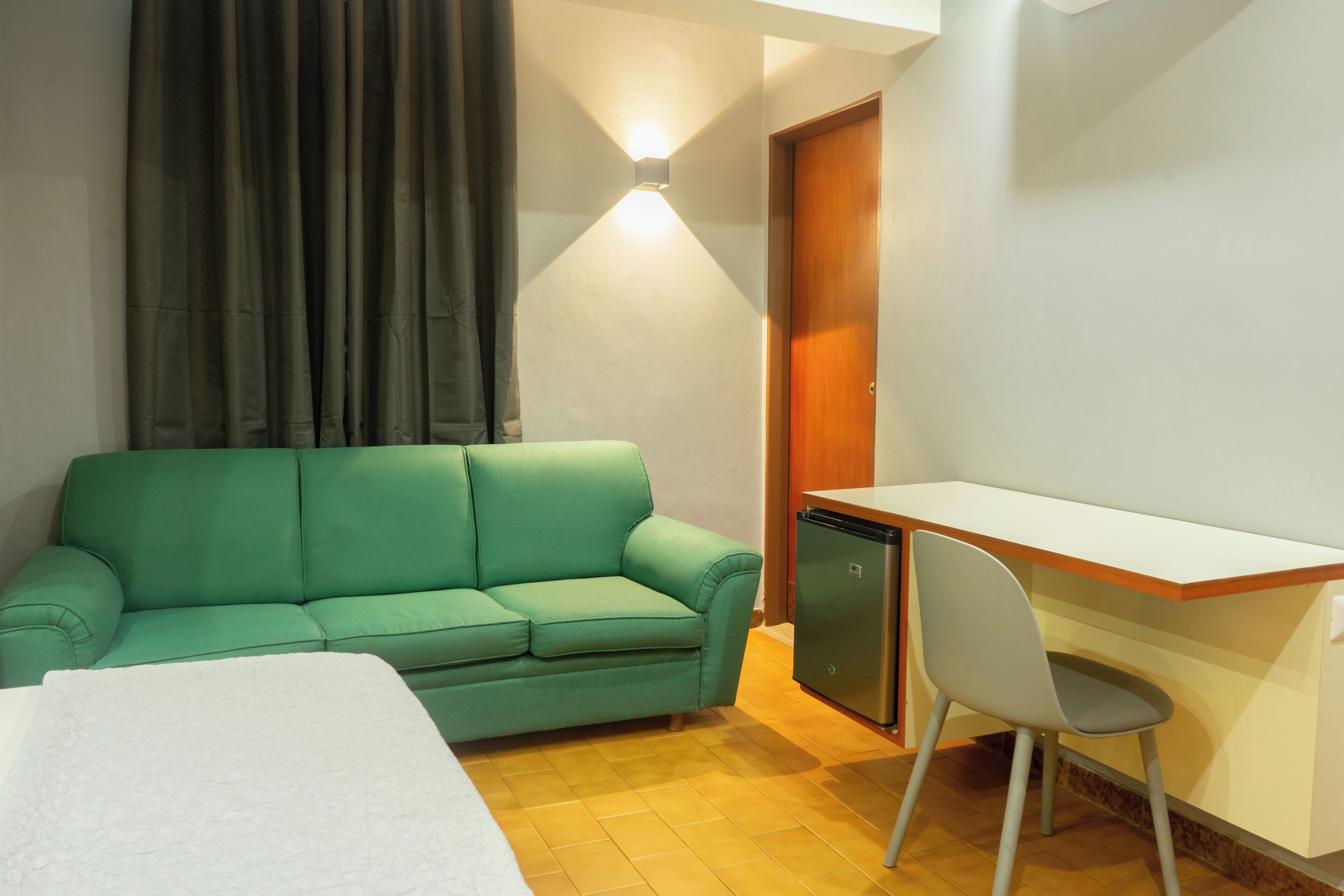 A room with a green sofa, a small desk with a chair, a white bed, and a door.